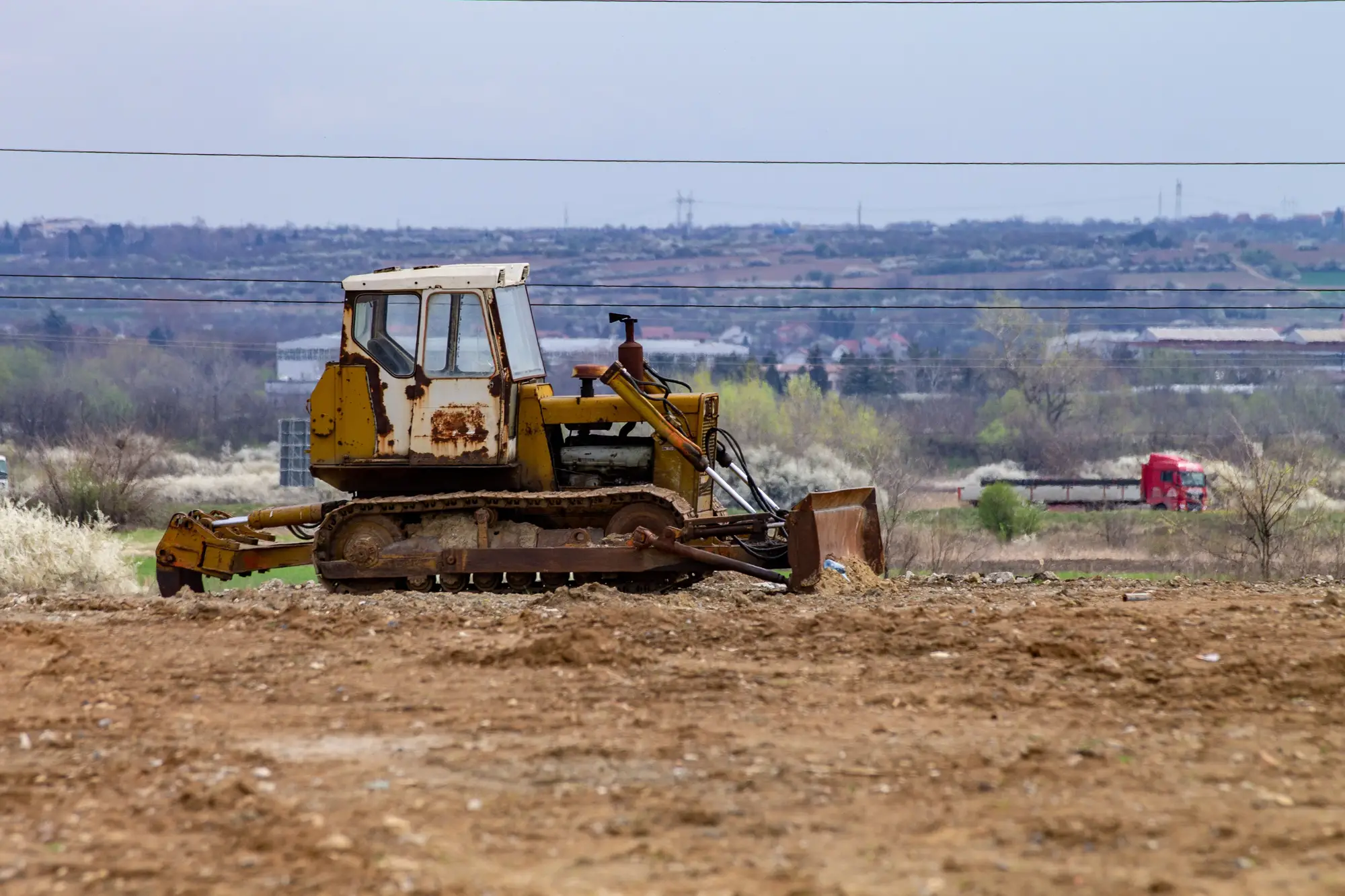 An old bulldozer parked on freshly scraped ground at the edge of a town, fields and power lines behind it